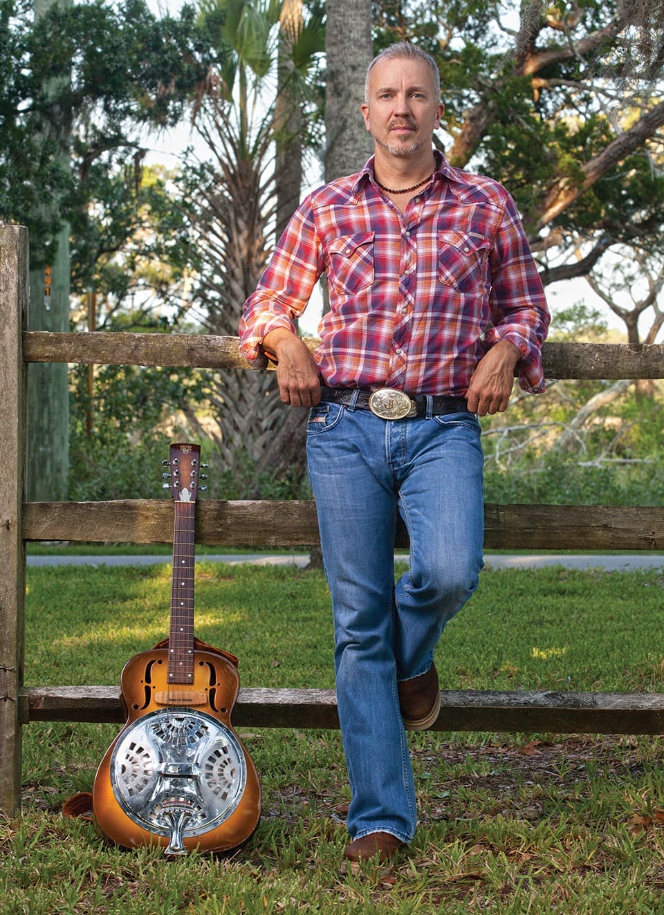 Red, White and JJ GREY 1 JJ Grey wears a red plaid shirt, a belt with a large buckle and jeans as he and his Dobro guitar rest against a wooden fence.